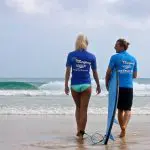 Two surfers in blue shirts on Byron Bay beach prepare for a 1.5-hour private surf lesson with Let's Go Surfing, watching the waves.