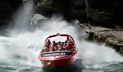 Experience the thrill as a red Shotover Jet boat filled with excited passengers races past Queenstown’s dramatic rocky cliffs, splashing water.