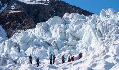 Tourists in winter gear trek across Franz Josef Glacier on a guided tour, with dramatic jagged ice formations and soaring alpine peaks behind.