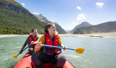 Two adventurers kayak a bright red Funyaks inflatable on the picturesque Dart River, framed by majestic mountains and lush forest, beneath a vibrant blue sky.
