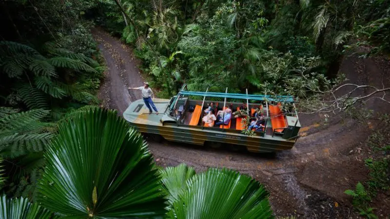 Express Kuranda Later Departure tour coach with excited passengers travels along a scenic dirt track surrounded by vibrant tropical rainforest.