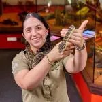 Smiling woman in khaki shirt holding a python at Cairns City Sights reptile exhibit, with glass animal enclosures in the background.