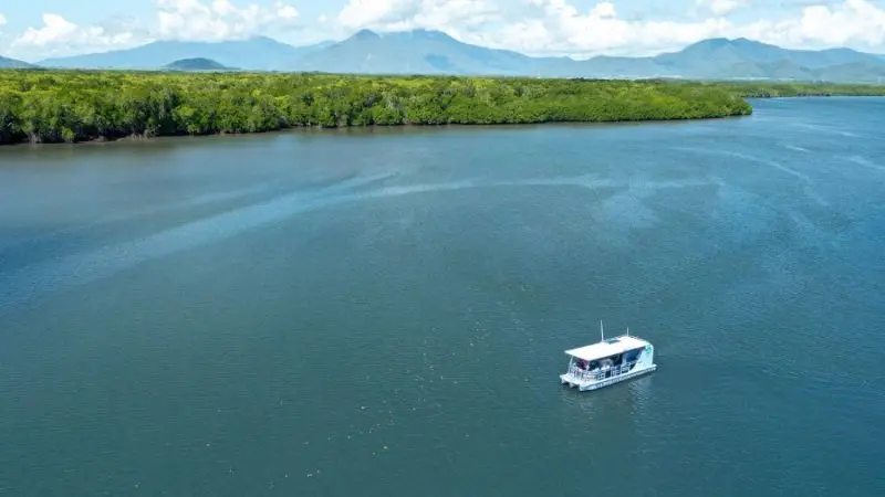 Experience a scenic Cairns City Sights River Cruise as a small white boat glides along a wide river, framed by lush greenery and mountains.