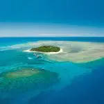 Discover a stunning, lush island near Cairns City Sights, encircled by turquoise waters and vibrant coral reefs beneath a sunny blue sky.