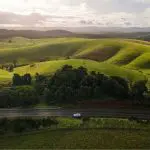 Sleek white car on a Private Exclusive Atherton Tablelands Tour cruises lush green hills beneath a dramatic sunrise or sunset sky.