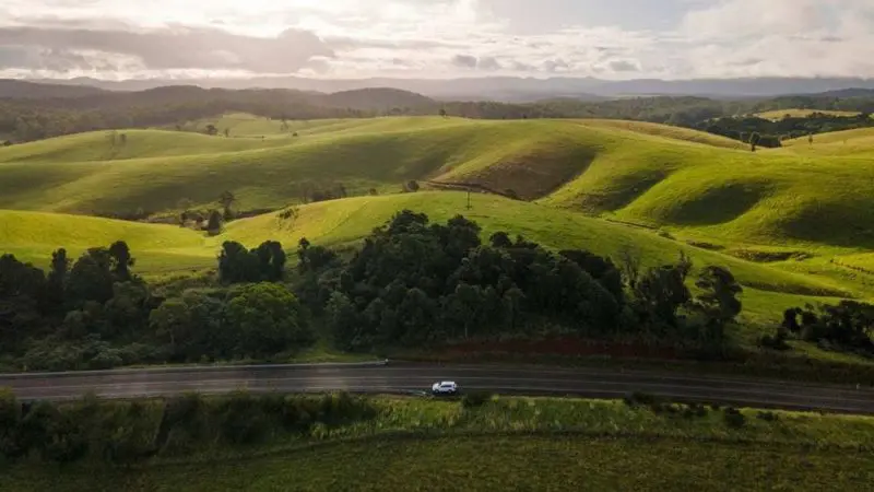 Sleek white car on a Private Exclusive Atherton Tablelands Tour cruises lush green hills beneath a dramatic sunrise or sunset sky.