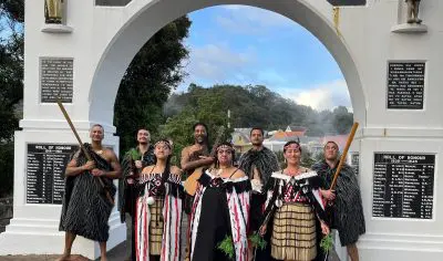Eight individuals in traditional Māori dress at Whakarewarewa stand before the “TEHOKOWHITU-A-TU” memorial archway.