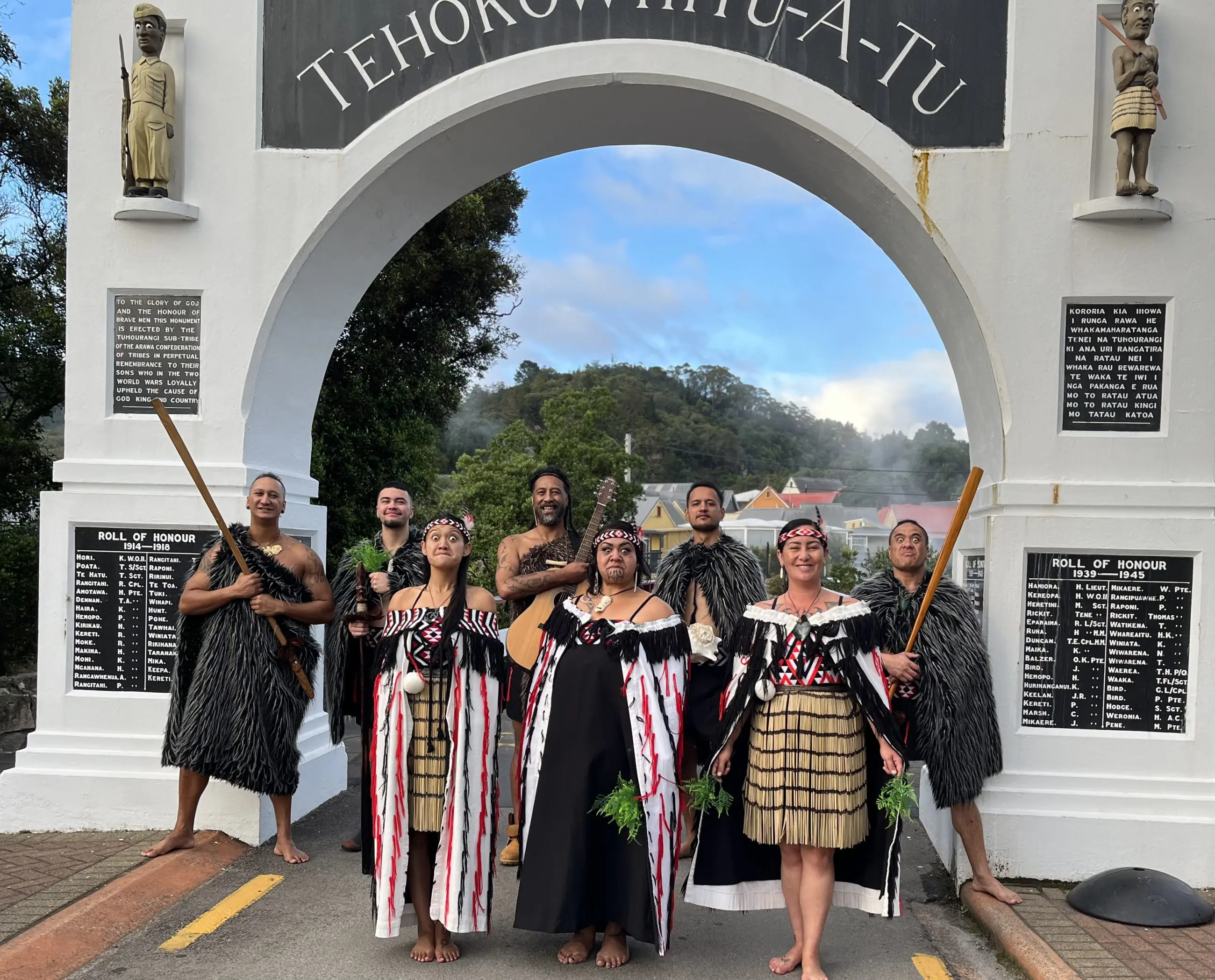 Eight performers in vibrant traditional Māori dress pose beneath a white archway after a Whakarewarewa Cultural Performance in New Zealand.