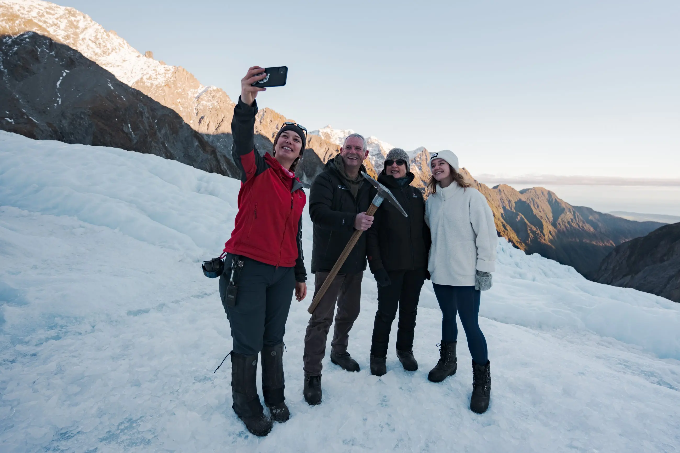Four friends in winter gear snap a selfie atop a snowy glacier, majestic mountain peaks and stunning alpine scenery in the background.