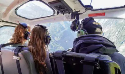 Three people with headsets experience an epic glacier helicopter tour, flying over snow-capped mountains under clear blue skies.