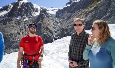 Three adventurers in outdoor gear smile on snowy ground during a Glacier Encounter, with sunlit rocky mountains in the background.