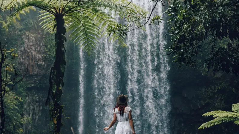 A woman in a flowing white dress stands beside a cascading waterfall, evoking the 16 Day Best Of East Coast Australia Comfort Tour experience.