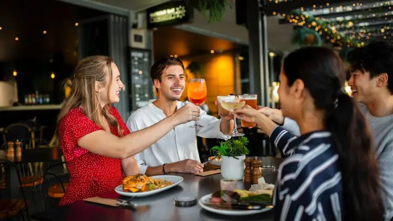 Four friends cheer with drinks at dinner, celebrating their 8-Day Sydney to Brisbane with Fraser Island Comfort Tour adventure.