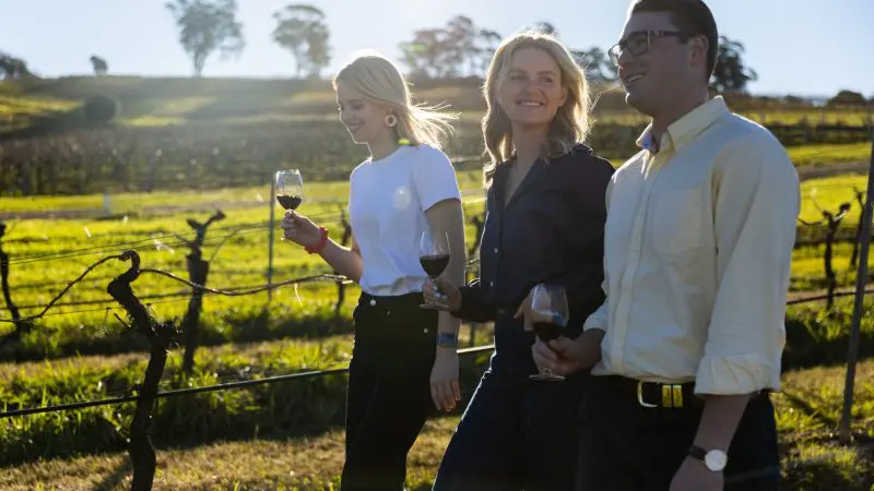 Three travellers enjoy wine whilst walking through a sunlit vineyard on their 14-day Sydney to Cairns Fraser Island Whitsundays tour.