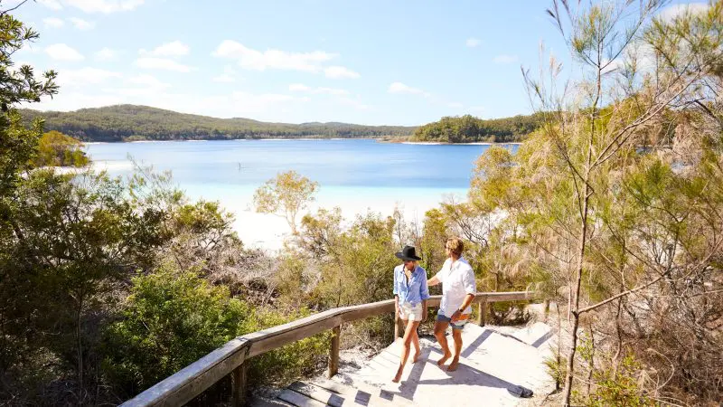 Two travellers hike sandy steps by lush trees, overlooking a blue lake and rolling hills on the 8-Day Sydney to Brisbane Fraser Island Tour.