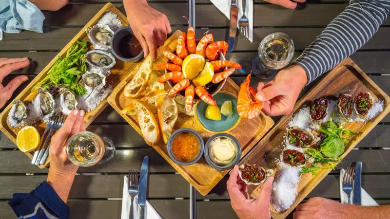 Three people enjoy seafood platters and drinks outdoors, savouring fresh coastal cuisine like a Perth to Exmouth Comfort Tour.