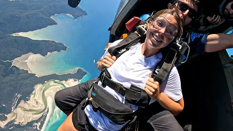 Excited woman in safety harness readies for tandem skydive at Whitehaven Beach, captured on video and photos above stunning coastline.