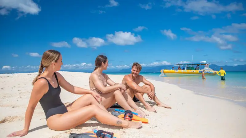 Three snorkellers chat on the sandy shore during a Great Barrier Reef Eco Adventure; tour boat anchored in crystal-clear water nearby.