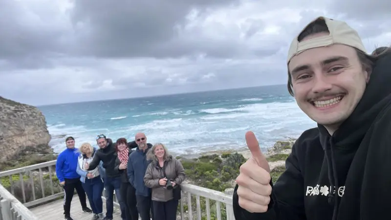 A happy person gives a thumbs up in front of the Kangaroo Island 2 Day Wildlife Adventure group by the sea under cloudy skies.