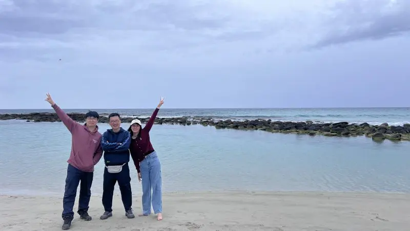 Three smiling people on Kangaroo Island’s sandy beach during a 2-Day Wildlife Adventure, with calm sea, rocky shore, and cloudy sky.
