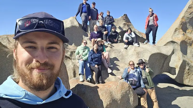 Excited group poses on rocky terrain during Kangaroo Island 2 Day Wildlife Adventure; man in hat snaps vibrant outdoor selfie.