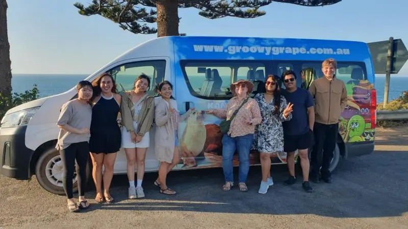 Group of eight excited travellers pose by Groovy Grape van on the beach, set for a Kangaroo Island 2 Day Wildlife Adventure tour.