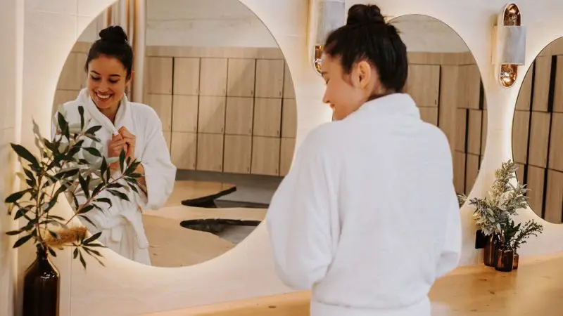 Smiling woman in a white dressing gown admires her reflection during a relaxing Peninsula Hot Springs Spa day, enjoying shuttle bus entry.