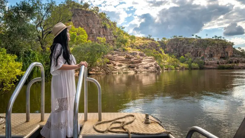Woman in a white dress takes a 1 Day Katherine Gorge Cruise, admiring dramatic rocky cliffs and lush trees beneath an overcast sky.