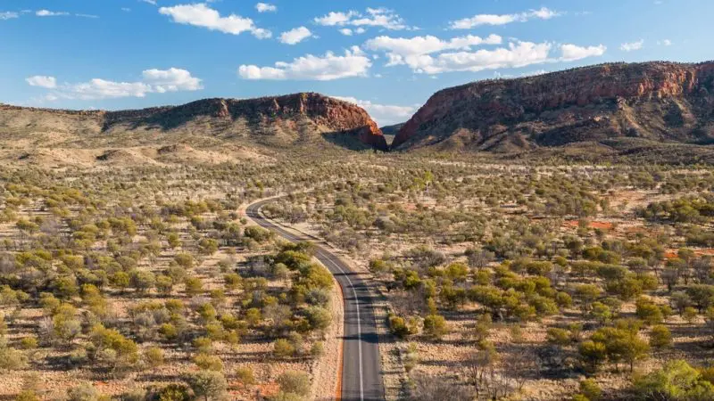 Scenic winding road through lush West MacDonnell Ranges to Standley Chasm, framed by a vivid blue sky dotted with white clouds.