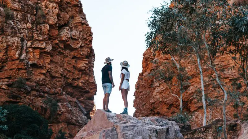 Two people in sun hats atop a rock, surrounded by striking red cliffs, trees, and gorges on a West MacDonnell Ranges tour.