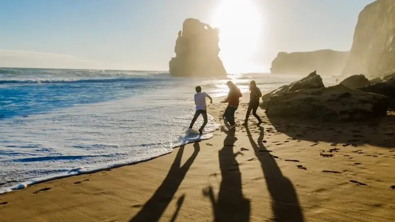 Three visitors jog along a golden beach at sunset, showcasing the scenic beauty of a top-rated Great Ocean Road Sunset Tour from Melbourne.