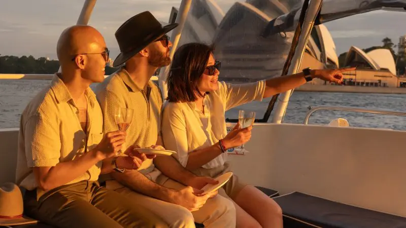 Three friends toast drinks on a BYO Sunset Sparkle Catamaran Cruise on Sydney Harbour, pointing to the iconic Opera House at sunset.