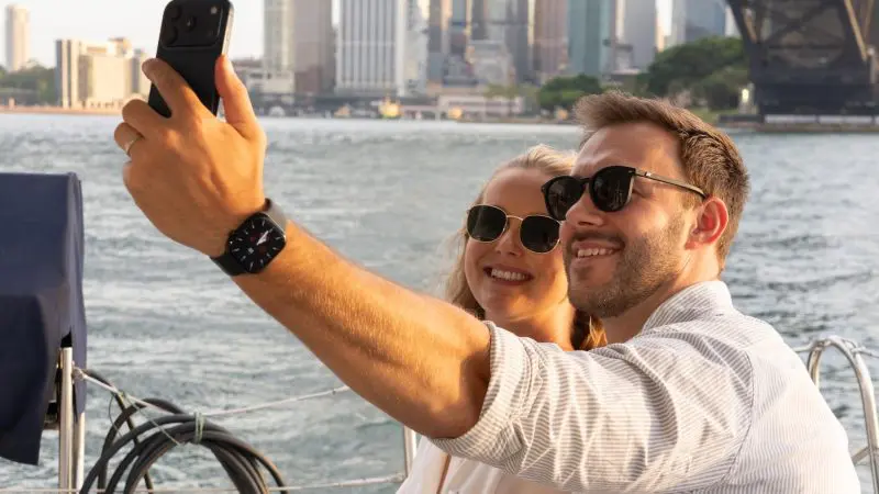 Smiling couple in sunglasses captures a selfie whilst enjoying a Sydney Harbour BYO Sunset Sparkle Catamaran Cruise at sunset.