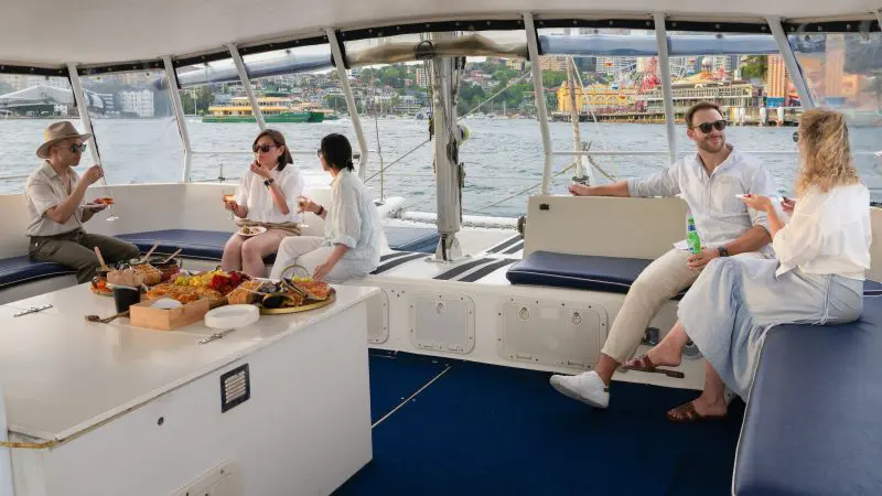 Group of friends enjoy food and drinks, laughing together on a Sydney Harbour BYO Sunset Sparkle Catamaran Cruise at dusk.