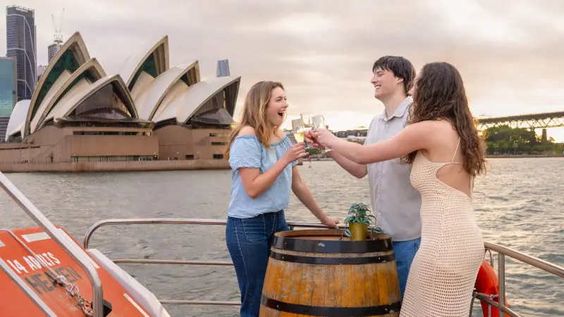 Three friends toast drinks on a Sydney Harbour sunset cruise near the Opera House, smiling and admiring the golden waterfront view.