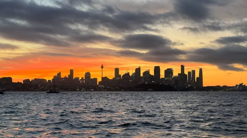 Scenic Sydney Harbour sunset cruise featuring stunning skyline views, vivid orange sky, dramatic clouds, and shimmering water reflections.