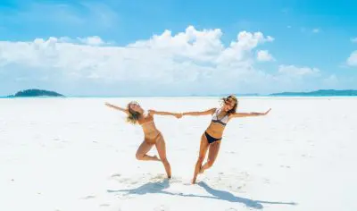 Two women in bikinis relax on a sunlit, sandy beach—ideal for an affordable 16-day holiday package at just £99 per day.
