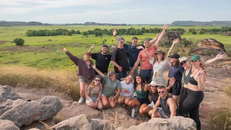 A smiling group on a 3 Day Kakadu Adventure Camping tour, posed on rocky terrain with lush green fields and scenic hills in the background.