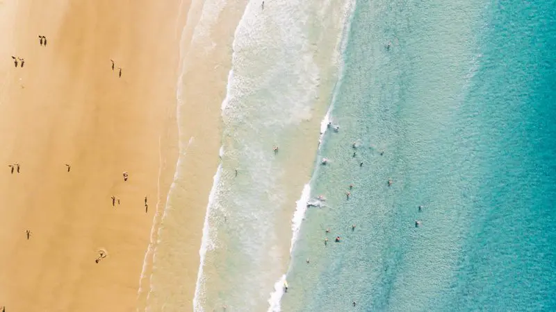 Aerial drone shot of beachgoers relaxing on golden sand, some swimming in crystal clear turquoise sea waves under bright sunny skies.