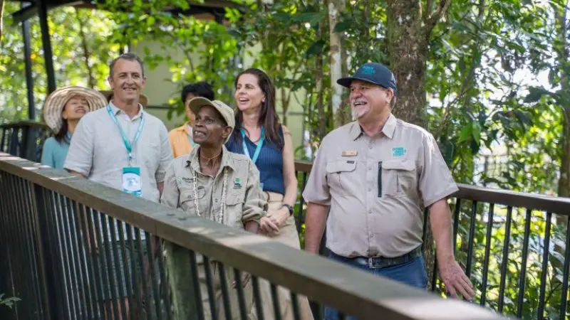 A group of people smiles whilst walking through a lush, green forest near a sparkling waterfall on a guided nature tour.