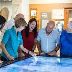 Six adults gather around an interactive touchscreen display during a CSHD Tour in a brightly lit Cairns City museum.