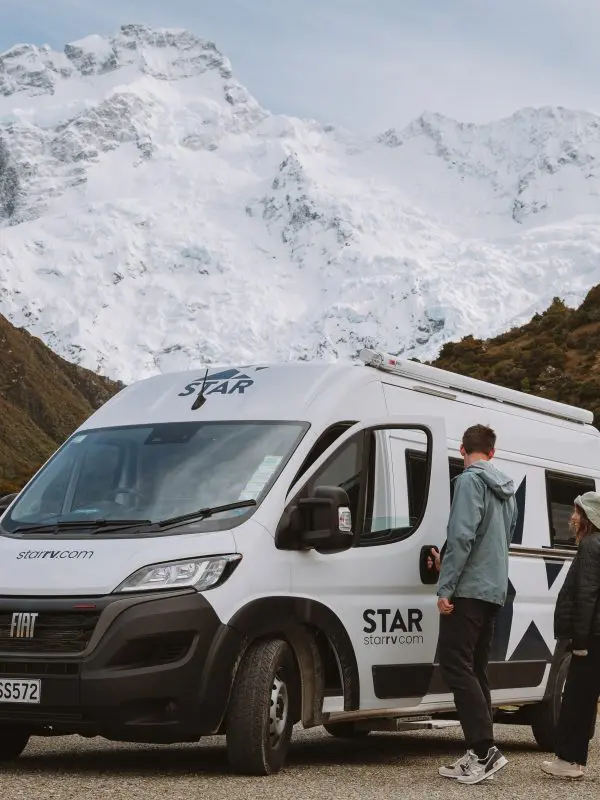 Two travellers stand beside a camper van, framed by majestic snow-capped mountains and lush green hills under a clear sky.