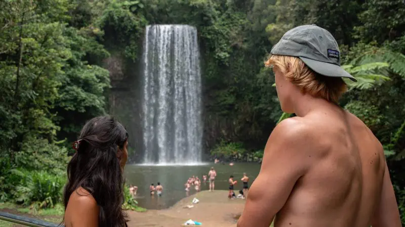 two friends standing in front of the millaa millaa waterfalls with people swimming in the plunge pools at the Atherton Tablelands