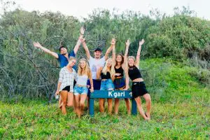 Eight happy young adults pose with arms up behind a Kgari sign in a lush, grassy forest, enjoying an outdoor group adventure on their Tag Along K'gari Fraser Tour