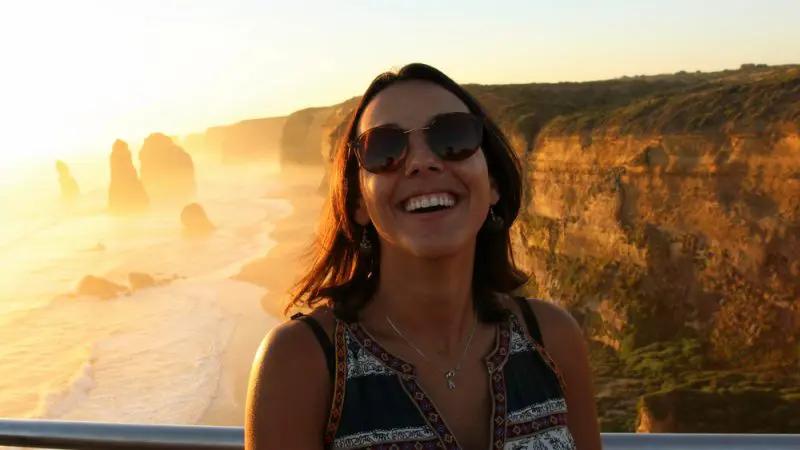 Smiling woman in trendy sunglasses stands by a scenic seaside cliff at sunset, with dramatic rock formations and ocean waves behind her.