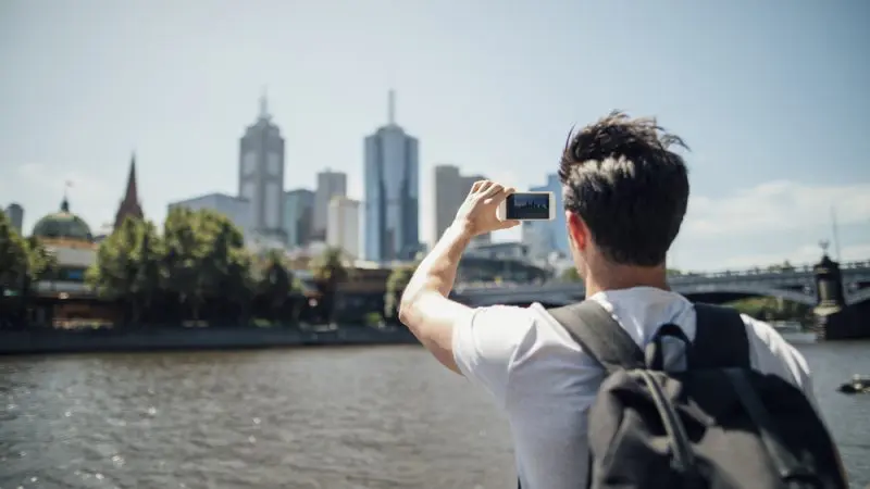 A tourist with a rucksack photographs a vibrant city skyline across a sparkling river under bright sunshine, capturing urban scenery.
