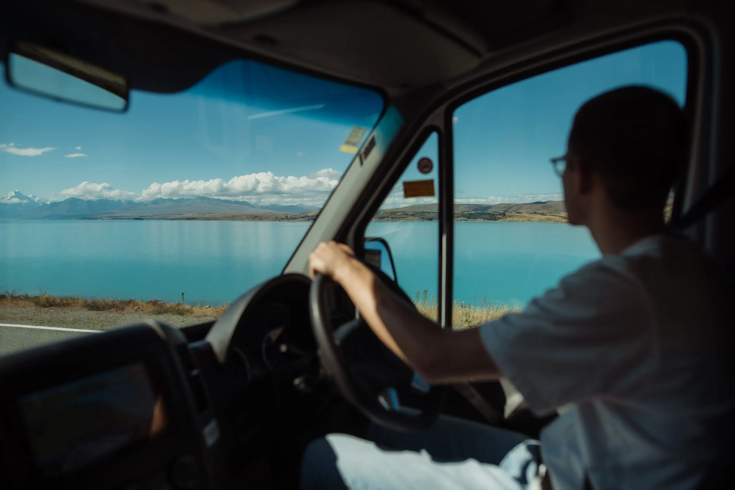 Driver in a van driving past lake tekapo with blue water and sky