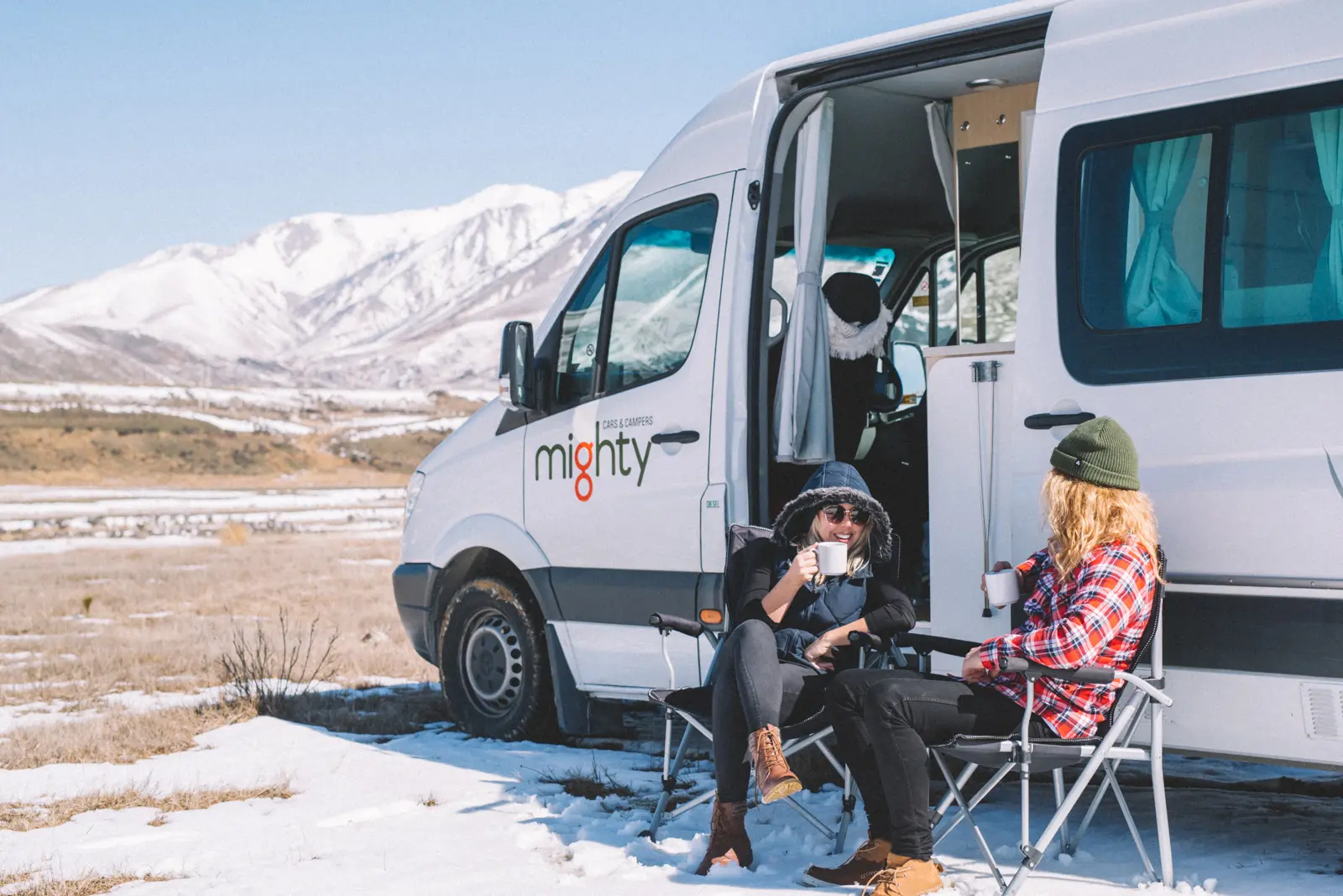 two people sitting outside of their van drinking coffee infront of snowy mountains