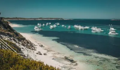 coastal beach with blue water and white sailboats in the daytime. the coastline has white sand and green bush shrubbing up a hill