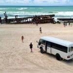 White coach parked on sandy Fraser Island beach by holidaymakers and iconic rusted shipwreck, K'gari Day Tour Ex Noosa, Australia.